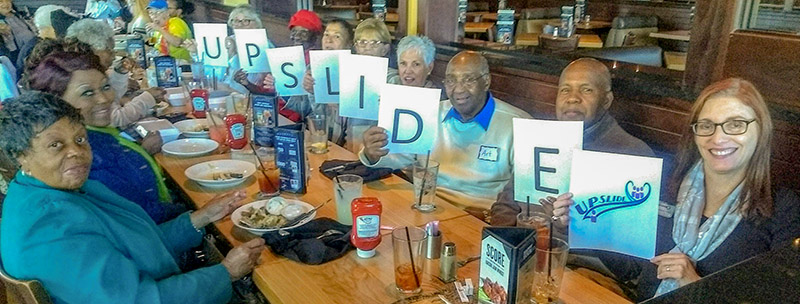 A group of seniors holding up pages that spell UPSLIDE, enjoying lunch together at a restaurant.